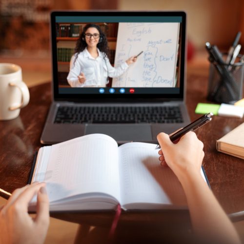 Online Course Concept. Point of view of female student sitting at table, writing in notebook, taking notes, using laptop, having online class with English teacher. Remote distant studying from home